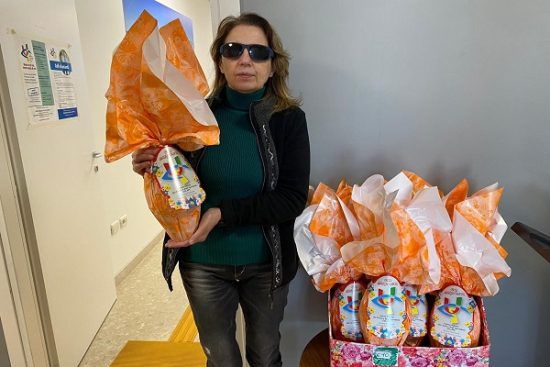 La nostra presidente Gigliola Chiappini posa con un un bell'uovo di Pasqua tra le mani. Indossa jeans scuri, una dolcevita verde bottiglia e un cardigan nero. L'uovo è confezionato in una allegra carta arancione e riporta il logo dell'Uici. In basso a destra un cesto pieno di altre uovo pronte per essere ordinate